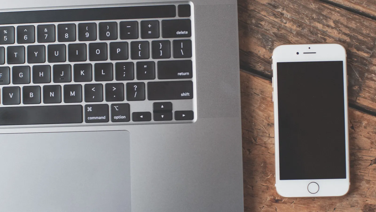 Top-down view of a silver laptop keyboard beside a white smartphone with a dark screen on a weathered wooden desk